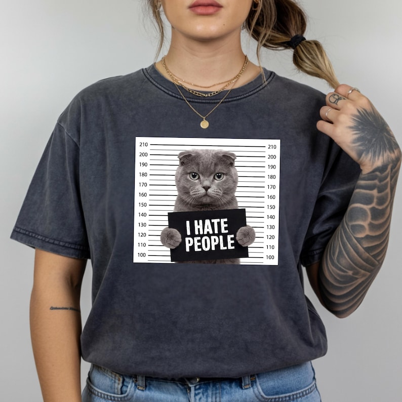 May include: Dark grey t-shirt featuring a grey cat holding a sign that reads "I HATE PEOPLE". The cat is positioned in front of a height chart. The shirt is a casual wear item.