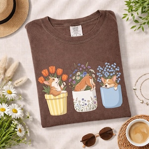 May include: Brown t-shirt featuring a graphic of three corgi puppies in flower pots with tulips, wildflowers, and forget-me-nots. The shirt has a Comfort Colors tag.