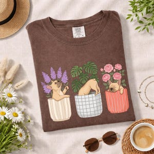 May include: Brown Comfort Colors t-shirt featuring a whimsical design of three French Bulldogs in decorative pots with plants and flowers. The shirt has a crew neck and a tag that says "Comfort Colors".