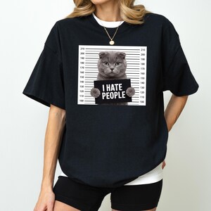 May include: Black t-shirt featuring a gray cat holding a sign that reads "I HATE PEOPLE" in white text. The cat is positioned in front of a height chart. The shirt is a casual style.