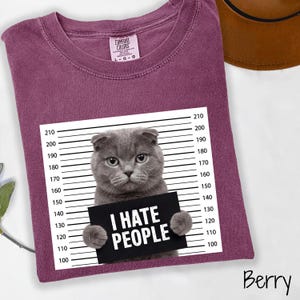 May include: Berry-colored t-shirt featuring a gray cat holding a sign that reads "I HATE PEOPLE." The cat is positioned in front of a height chart. The shirt has a crew neck and short sleeves. The brand label is visible on the upper left.