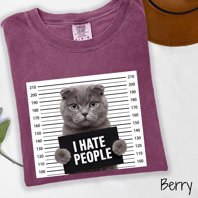 May include: Berry-colored t-shirt featuring a gray cat holding a sign that reads "I HATE PEOPLE." The cat is positioned in front of a height chart. The shirt has a crew neck and short sleeves. The brand label is visible on the upper left.