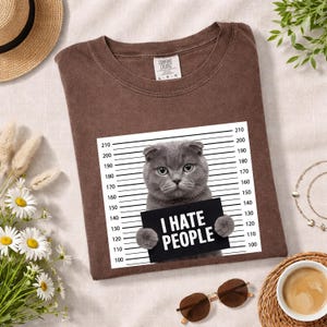 May include: Brown t-shirt featuring a gray cat holding a sign that reads "I HATE PEOPLE." The cat is positioned in front of a height chart. The shirt has a crew neck and short sleeves.