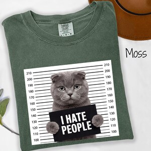 May include: Moss green t-shirt with a graphic of a gray cat holding a sign that reads "I HATE PEOPLE". The shirt has a crew neck and short sleeves. The cat is positioned in front of a police lineup background.