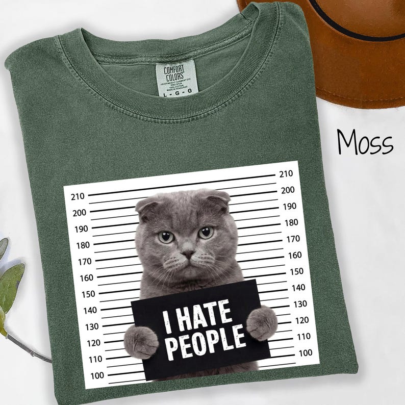 May include: Moss green t-shirt with a graphic of a gray cat holding a sign that reads "I HATE PEOPLE". The shirt has a crew neck and short sleeves. The cat is positioned in front of a police lineup background.