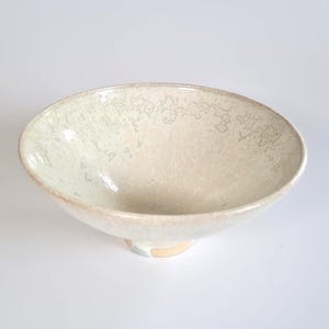 May include: A ceramic bowl with a wide, shallow basin and a tapered foot. The interior and exterior feature a glossy, off-white glaze with a subtle, intricate crackle pattern throughout. The rim is smooth, and the base shows a hint of light clay.