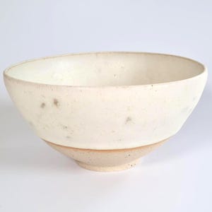 May include: A ceramic bowl with a wide, open rim and a tapered base. The upper portion features a smooth, off-white glaze with subtle grey speckles, while the lower section and base remain in an unglazed, textured, light tan clay finish.