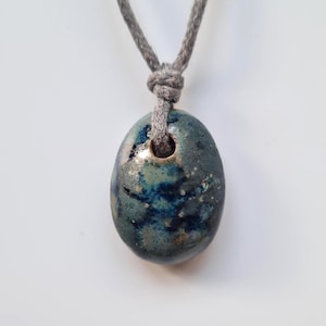 May include: Oval-shaped ceramic pendant with a blue and green glaze, suspended from a gray cord. The pendant has a hole at the top for the cord. The ceramic has a speckled appearance.