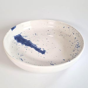 May include: A round ceramic bowl with a white base and a glossy finish. The interior features a dark blue paint splatter pattern, including a prominent elongated blue streak on the left side and smaller scattered blue dots across the surface.