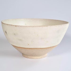 May include: A ceramic bowl with a wide, rounded rim and a tapered base. The upper portion features an off-white, matte glaze with subtle dark speckles, while the lower section remains an unglazed, light beige clay with a visible speckled texture.
