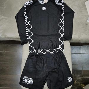 Könnte beinhalten: Schwarzes Sport-Langarmshirt und Shorts-Set. Das Shirt hat weiße Akzente und Text auf den Ärmeln, mit einem passenden Design auf den Shorts. Ein Logo ist auf Brust und Shorts sichtbar.