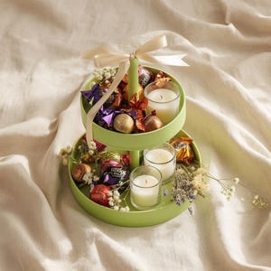 Könnte beinhalten: Ein zweistöckiges, hellgrünes Serviertablett mit verpackten Pralinen, kleinen weißen Kerzen und Trockenblumen. Oben ist eine cremefarbene Schleife gebunden. Das Tablett steht auf cremefarbenem Stoff.