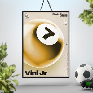 May include: A framed poster with a black border, featuring a gold and white abstract design with the text "Vini Jr." and "Rio de Janeiro Madrid". A soccer ball, cleats, and a trophy are in the foreground.