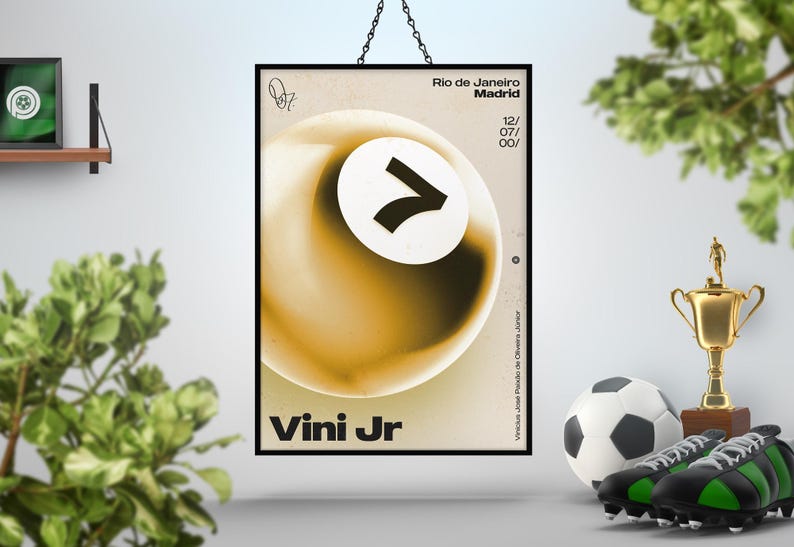 May include: A framed poster with a black border, featuring a gold and white abstract design with the text "Vini Jr." and "Rio de Janeiro Madrid". A soccer ball, cleats, and a trophy are in the foreground.