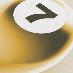 May include: Abstract art piece featuring a white circle with a black number seven. The background has a gradient of yellow and brown tones, with a textured appearance. The artwork has a modern, minimalist aesthetic.