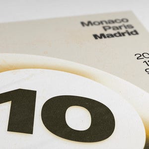 May include: A close-up of a beige and white poster with the number "10" in black on a white circle. The poster also features the words "Monaco Paris Madrid" and the numbers "20/12/981" in black.