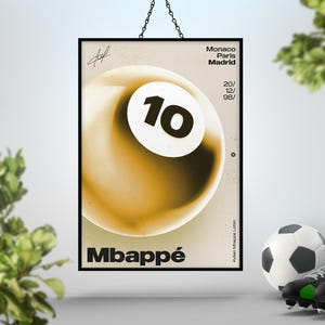 May include: A framed poster featuring a stylized image of a soccer ball with the number 10 on it. The poster has the name "Mbapp&eacute;" at the bottom and text that reads "Monaco Paris Madrid 20/ 12/ 98/". A soccer ball, cleats, and a trophy are in the foreground.