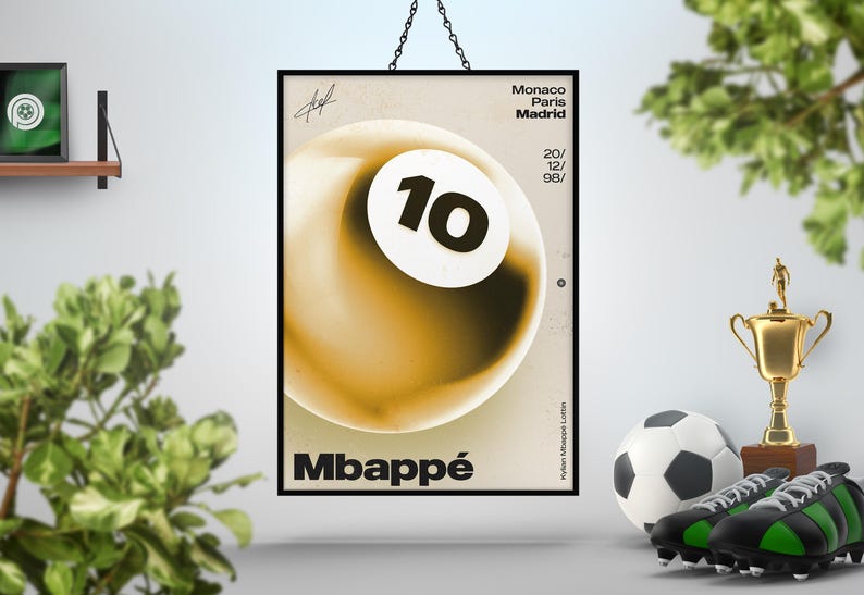 May include: A framed poster featuring a stylized image of a soccer ball with the number 10 on it. The poster has the name "Mbapp&eacute;" at the bottom and text that reads "Monaco Paris Madrid 20/ 12/ 98/". A soccer ball, cleats, and a trophy are in the foreground.