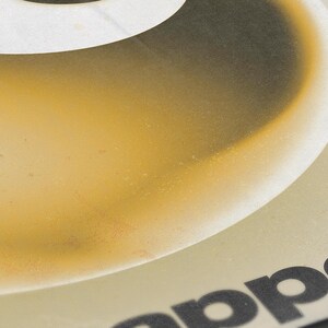 May include: Close-up of a poster with a gradient of yellow and brown colors. The top portion is white with a large black letter. The word "app&eacute;" is visible in black at the bottom right corner. The poster has a vintage aesthetic.