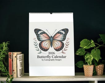May include: A 2026 Butterfly Calendar by Lunography Images. The calendar features a watercolor illustration of a butterfly with pink, blue, and brown wings. The calendar is white with a spiral binding and is displayed on a white shelf.