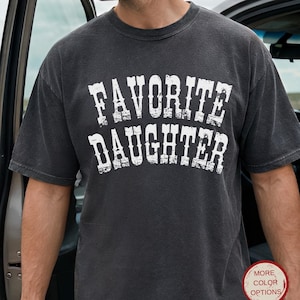 May include: A charcoal gray short-sleeve t-shirt with a crew neckline. The front features the text "FAVORITE DAUGHTER" printed in a distressed white font. A small circular badge in the lower right corner contains the text "MORE COLOR OPTIONS".