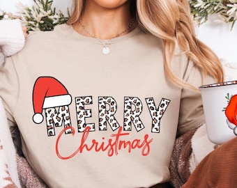 May include: Beige t-shirt with the words "MERRY Christmas" in a festive design. The word "MERRY" has a leopard print pattern, and the Santa hat is red. A white mug with a Santa Claus graphic is held in the hand.