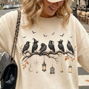 May include: A beige crewneck sweatshirt featuring a graphic print of six black crows perched on a branch, with hanging elements including a lantern, a spiderweb, and a tombstone. The sweatshirt is paired with a beige baseball cap and a black quilted bag.