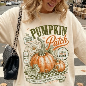May include: A beige crewneck sweatshirt featuring a graphic print of pumpkins with the text PUMPKIN PATCH, LOCALLY GROWN, FARM FRESH, and FRESH FROM THE PATCH, paired with a beige baseball cap with a small black logo and a black quilted shoulder bag.