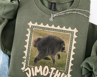 May include: An olive green crewneck sweatshirt featuring a graphic of a raccoon inside a postage stamp border. Below the stamp, the text "JIMOTHY" is printed in a light tan, rounded font. The sweatshirt has long sleeves and a ribbed collar.