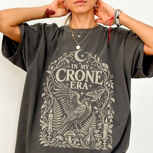 May include: A charcoal gray short-sleeve t-shirt featuring a cream-colored graphic print. The design includes a central raven, ferns, mushrooms, candles, a crescent moon, and the text "IN MY CRONE ERA" framed by intricate botanical illustrations.