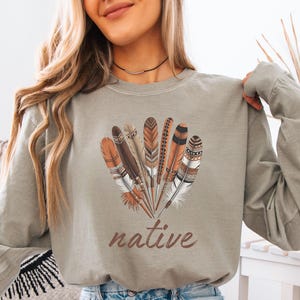 May include: A long-sleeved, olive green sweatshirt featuring a cluster of detailed feathers in shades of brown, orange, and white. The word "native" is printed in a brown script font below the feathers. The shirt is made of a soft material.