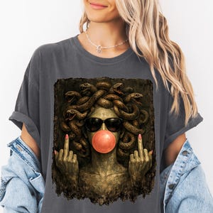 May include: A charcoal gray t-shirt featuring a graphic print of a Medusa figure with snakes for hair, wearing black sunglasses and blowing a pink bubblegum bubble. The figure is shown with both middle fingers raised in a defiant gesture.