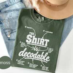 May include: A moss green t-shirt with white text reading The Shirt is decodable, surrounded by linguistic labels like Digraphs and r-controlled. The shirt is layered with light blue denim jeans, a tan wide-brimmed hat, and a gold chain necklace.