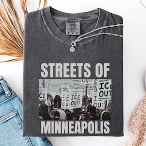 May include: Dark gray t-shirt with the text "STREETS OF MINNEAPOLIS" in white. The shirt features a black and white image of a protest. A silver necklace with a pendant is on the shirt. The shirt is folded on a white surface.