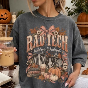 May include: A charcoal gray short-sleeve t-shirt featuring a graphic print with the text RAD TECH radiologic technologist. The design includes illustrations of an X-ray, a skeleton hand, a skull, pumpkins, books, and a pink ribbon bow.
