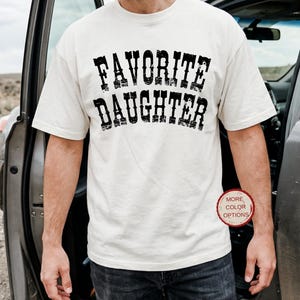May include: A cream-colored short-sleeve t-shirt featuring the text FAVORITE DAUGHTER printed in a distressed black font across the chest. The shirt has a crew neckline and a standard fit, with a small circular red badge stating MORE COLOR OPTIONS.