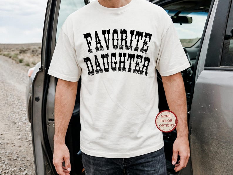 May include: A cream-colored short-sleeve t-shirt featuring the text FAVORITE DAUGHTER printed in a distressed black font across the chest. The shirt has a crew neckline and a standard fit, with a small circular red badge stating MORE COLOR OPTIONS.