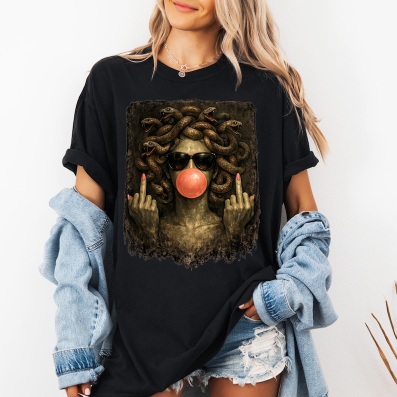 May include: A black t-shirt featuring a graphic print of a Medusa figure with snakes for hair, wearing black sunglasses and blowing a pink bubblegum bubble. The figure is shown with both middle fingers raised in a gesture against a dark background.