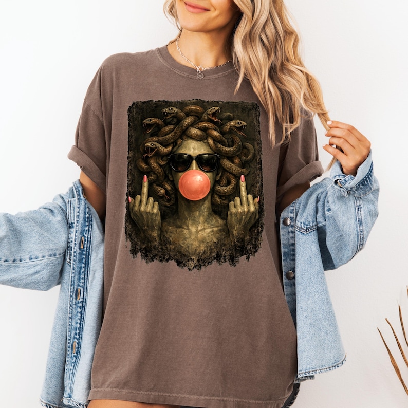 May include: A brown short-sleeve t-shirt featuring a graphic print of a figure with snakes for hair, wearing black sunglasses, blowing a pink bubblegum bubble, and holding up both middle fingers in a defiant gesture against a dark background.