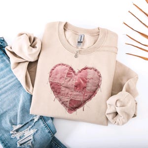 May include: A beige sweatshirt featuring a patchwork heart design in shades of pink and red. The heart is made of stitched fabric squares. A silver necklace with a small pendant is on the neckline. The sweatshirt is paired with blue jeans.