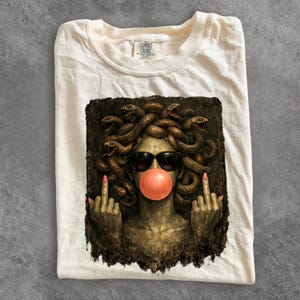 May include: A cream-colored t-shirt featuring a graphic print of a stone-textured figure with snakes for hair, wearing black sunglasses and blowing a pink bubblegum bubble. The figure has both hands raised with middle fingers extended.