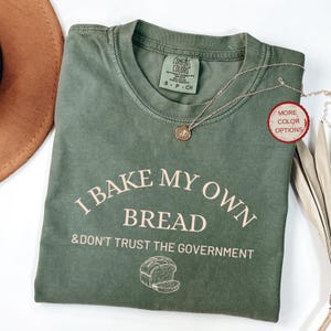 May include: Olive green t-shirt with the text "I BAKE MY OWN BREAD & DON'T TRUST THE GOVERNMENT" in a light beige color. A silver necklace with a round pendant is on the shirt. The shirt is folded on a white surface.