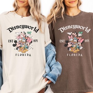 Puede incluir: Dos camisetas, una crema y otra marr&oacute;n, con un gr&aacute;fico de Disneyworld. El gr&aacute;fico incluye a Mickey y Minnie Mouse, Donald y Daisy Duck, Goofy y Pluto. El texto dice "Disneyworld EST 1971 FLORIDA".