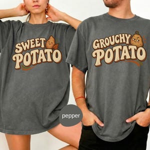 May include: Two charcoal gray t-shirts featuring graphic prints. The left shirt displays the text "SWEET POTATO" with a smiling potato illustration. The right shirt displays the text "GROUCHY POTATO" with a frowning potato illustration.