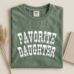 May include: A sage green short-sleeved t-shirt with a crew neckline. The shirt features the white distressed text "FAVORITE DAUGHTER" printed in a bold, arched font across the chest. The garment includes a Comfort Colors brand neck label.