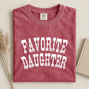 May include: A crimson red short-sleeved crew neck t-shirt featuring the white distressed text "FAVORITE DAUGHTER" printed in a bold, arched font across the chest. The garment includes a Comfort Colors brand label inside the neckline.