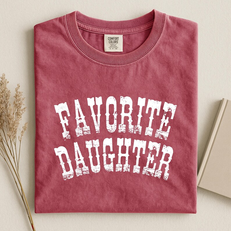 May include: A crimson red short-sleeved crew neck t-shirt featuring the white distressed text "FAVORITE DAUGHTER" printed in a bold, arched font across the chest. The garment includes a Comfort Colors brand label inside the neckline.