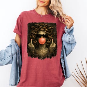 May include: A faded red short-sleeve t-shirt featuring a graphic print of a Medusa figure with snakes for hair, wearing black sunglasses, blowing a pink bubblegum bubble, and holding up both middle fingers in a defiant gesture.