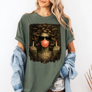 May include: An olive green t-shirt featuring a graphic print of a Medusa figure with snakes for hair, wearing black sunglasses and blowing a pink bubblegum bubble. The figure is shown with both middle fingers raised in a gesture against a dark background.