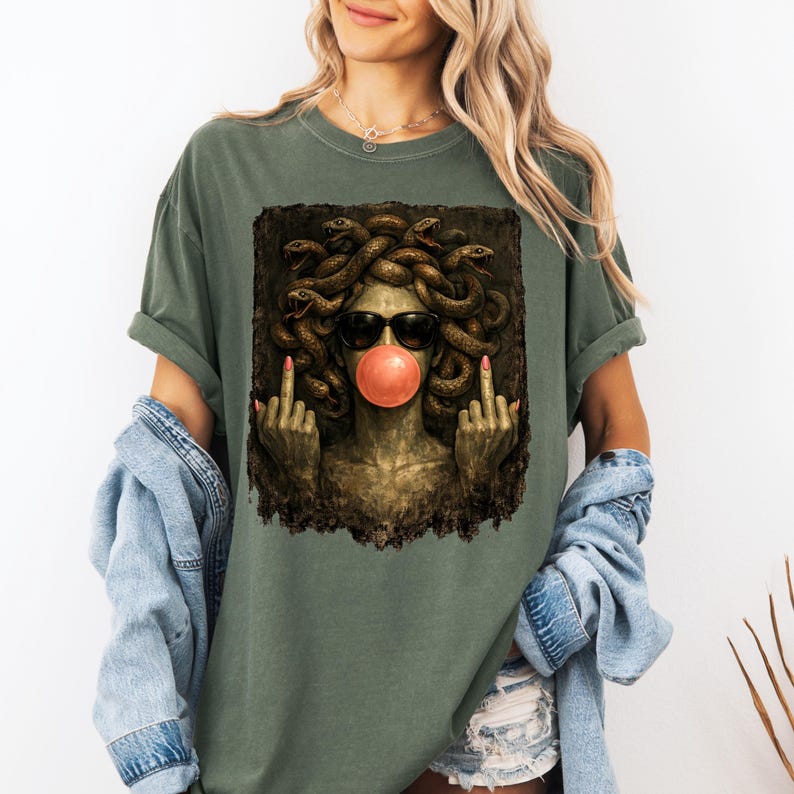 May include: An olive green t-shirt featuring a graphic print of a Medusa figure with snakes for hair, wearing black sunglasses and blowing a pink bubblegum bubble. The figure is shown with both middle fingers raised in a gesture against a dark background.